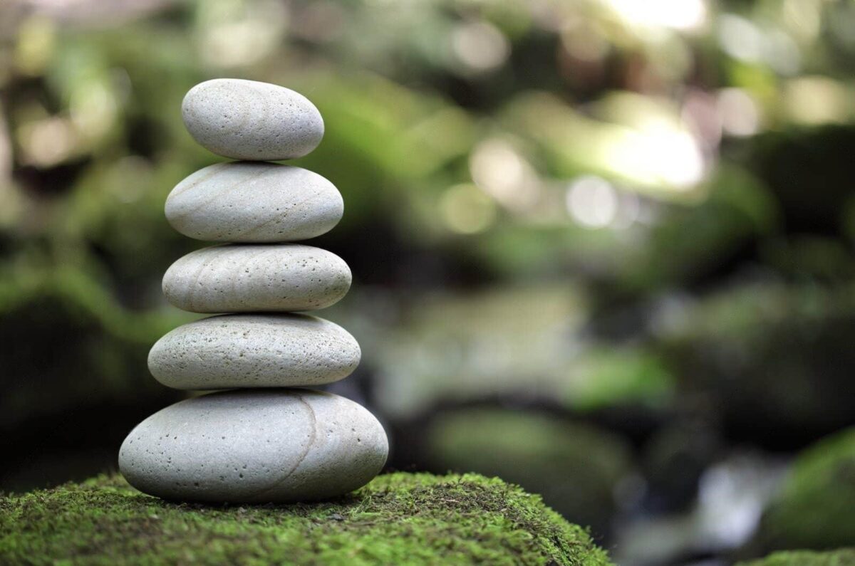 “A stack of six smooth, round stones of varying sizes balanced on top of each other in ascending order. The stones are placed on a green mossy surface with a blurred background featuring what appears to be a natural environment with foliage.”